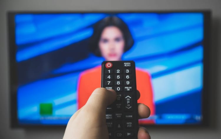 Nærbilde av svart fjernkontroll til tv. Litt uklart i bakgrunnen er det en tv-skjerm med en kvinne med rød jakke.
