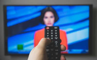 Nærbilde av svart fjernkontroll til tv. Litt uklart i bakgrunnen er det en tv-skjerm med en kvinne med rød jakke.