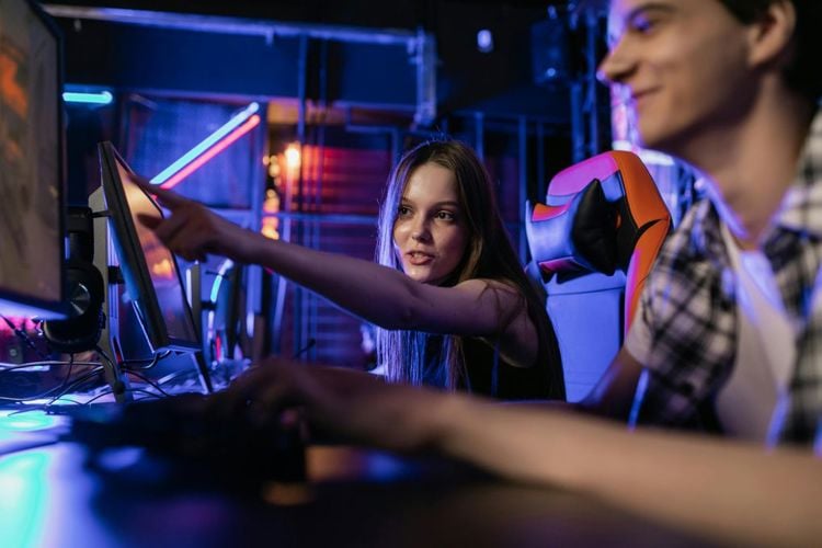 To tenåringer, en jente og en gutt, spiller dataspill sammen. Hun peker på skjermen foran dem og han smiler.