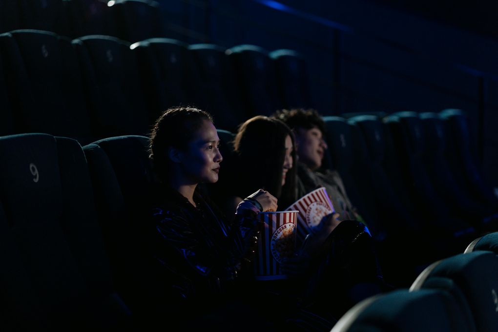 Tre personer sitter i en mørk kinosal. De sitter ved siden av hverandre,og har popcornbeger i hendene.