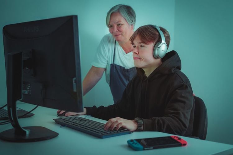 En tenåringsgutt sitter foran PC mens en voksen kvinne står ved siden av og følger med på skjermen. De er fotografert skrått forfra, slik at det ikke vises hva de ser på skjermen.