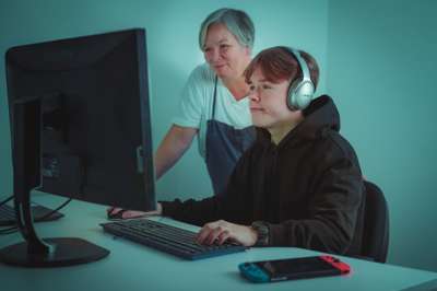En tenåringsgutt sitter foran PC mens en voksen kvinne står ved siden av og følger med på skjermen. De er fotografert skrått forfra, slik at det ikke vises hva de ser på skjermen.