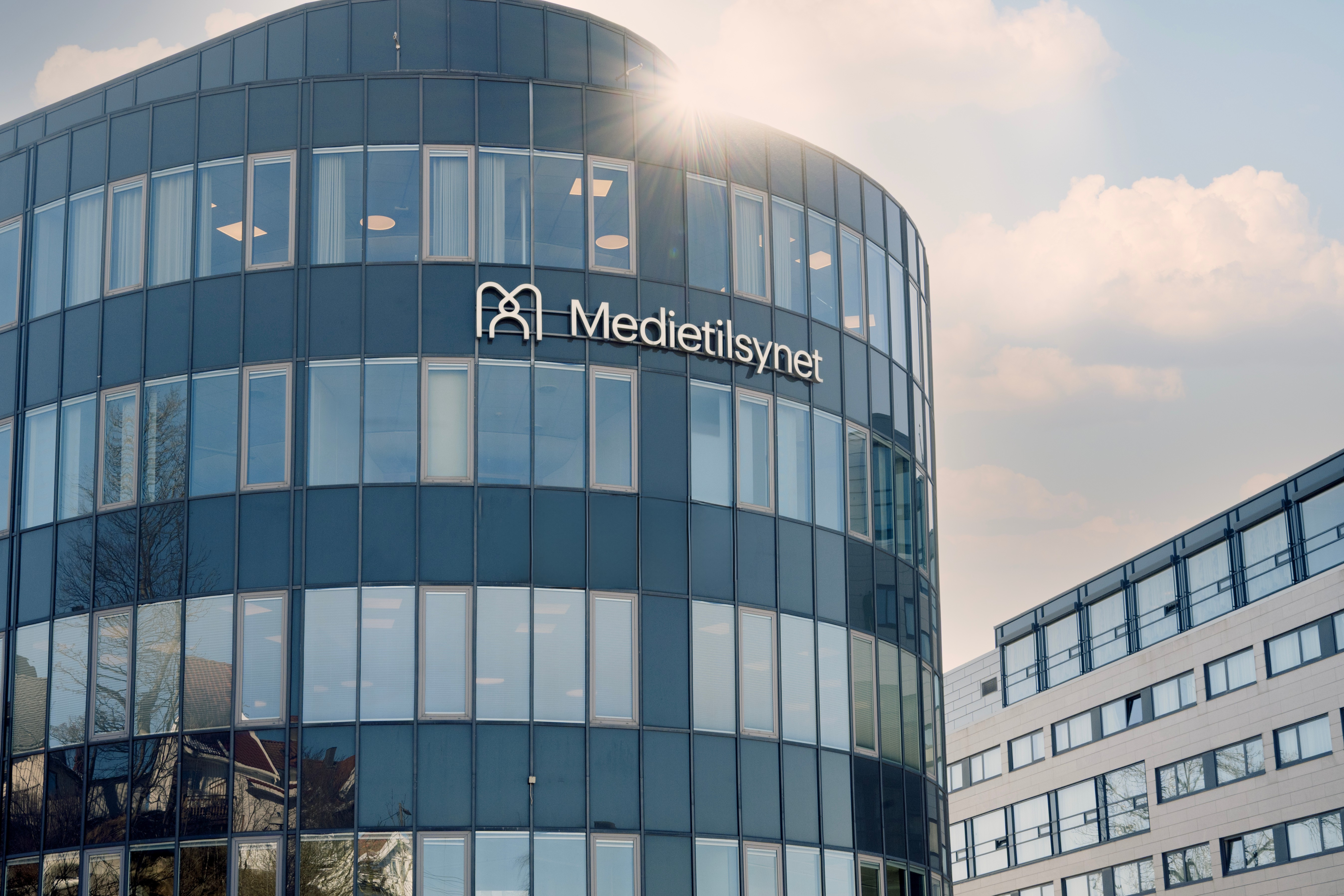Moderne kontorbygg med buet glassfasade og skiltet «Medietilsynet», sett i dagslys med sol som skinner over bygget.