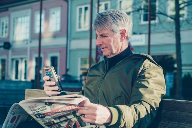 Mann leser aviser på mobil og papir