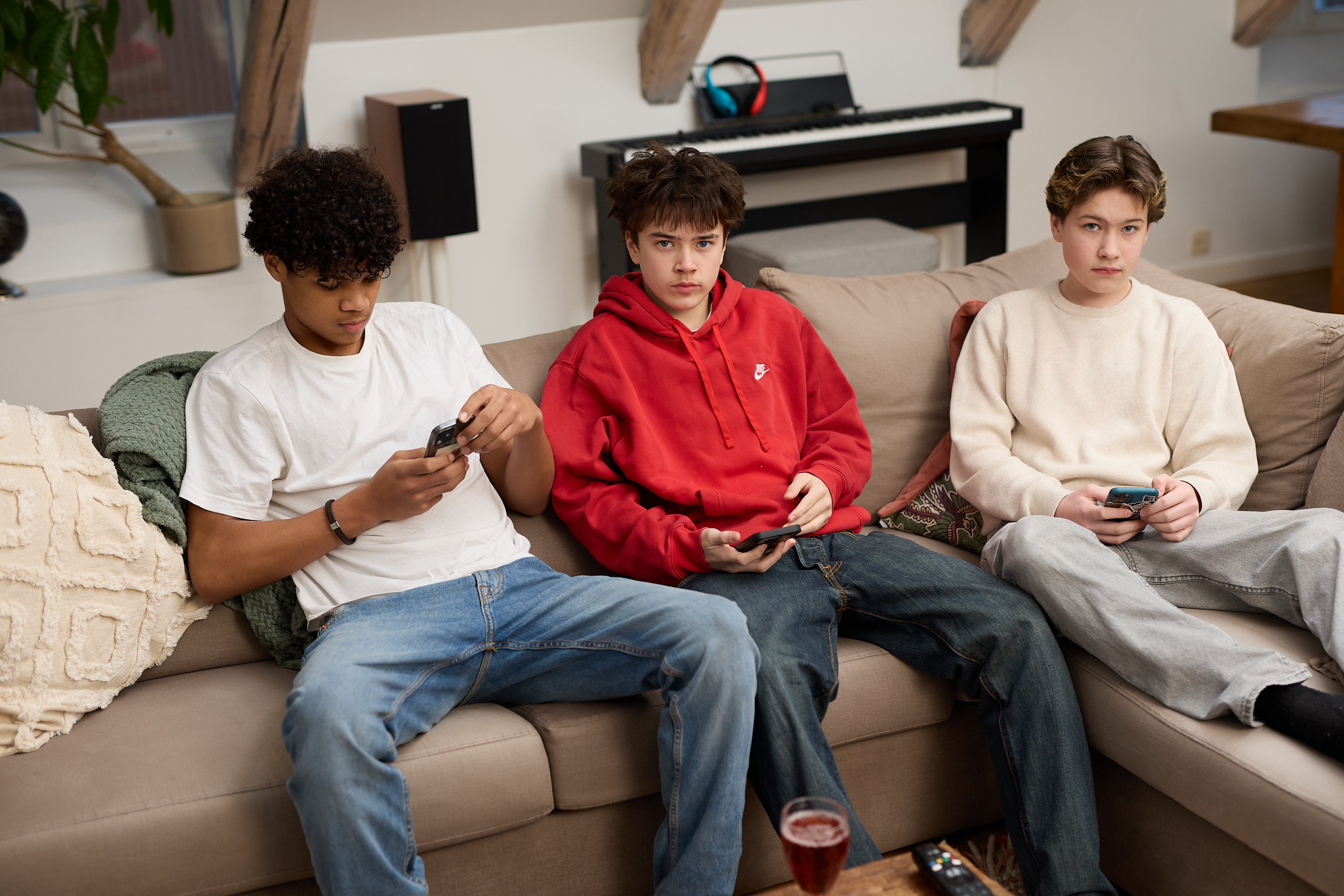Tre tenåringsgutter sitter i en beige sofa i en stue og ser på hver sin mobiltelefon. Gutten til venstre har hvit t-skjorte og jeans, gutten i midten har rød hettegenser, og gutten til høyre har lys genser og grå bukse. I bakgrunnen står et elektrisk piano med hodetelefoner på, og det ligger en fjernkontroll og et glass med rød drikke på bordet foran dem.