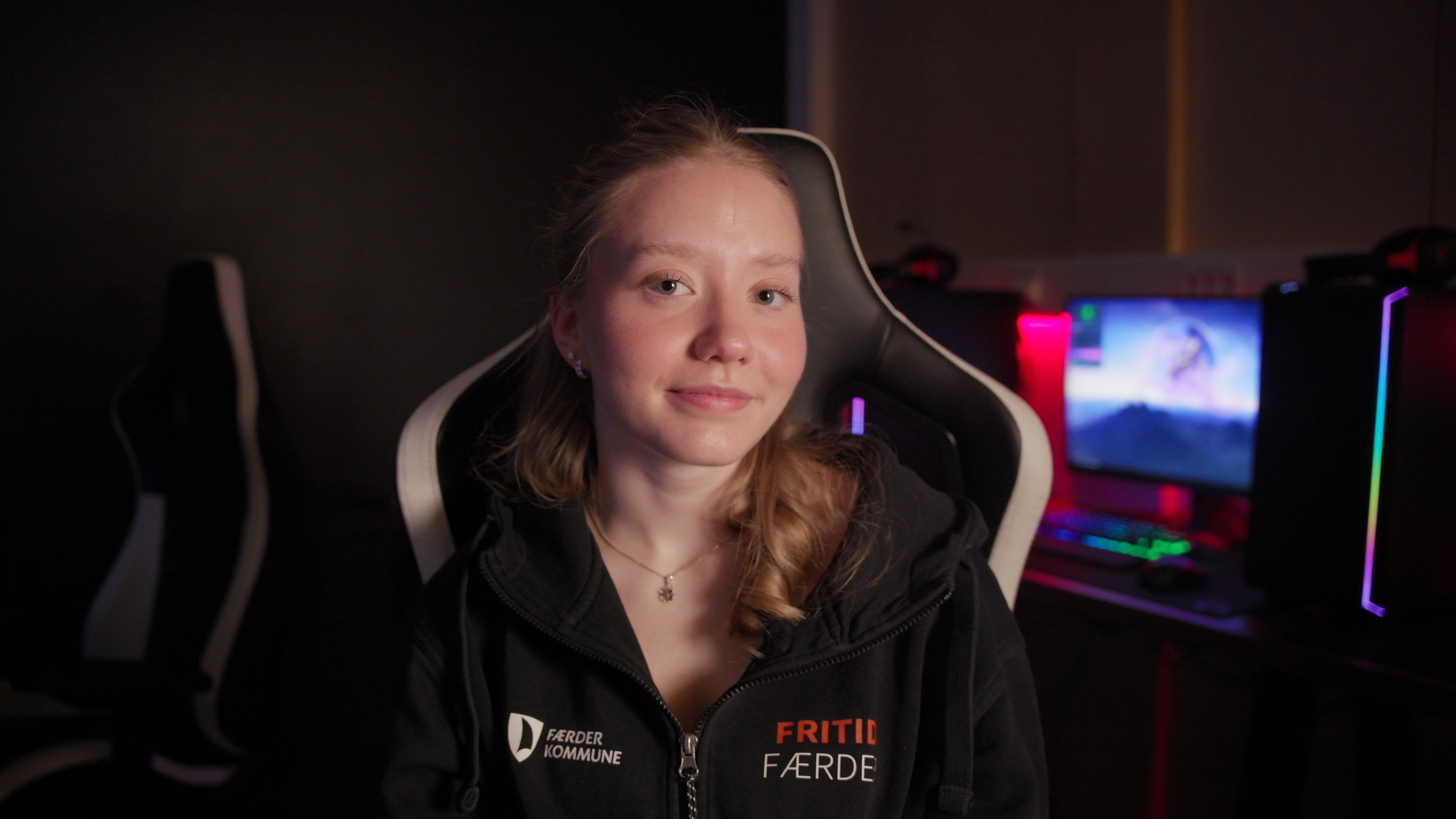 Ung kvinne sitter i en gamingstol i et mørkt rom med datamaskiner i bakgrunnen. Hun har på seg en hettegenser med teksten «Fritid Færder» og «Færder kommune».