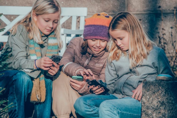 Tre jenter sitter på en benk og ser på mobiltelefoner.