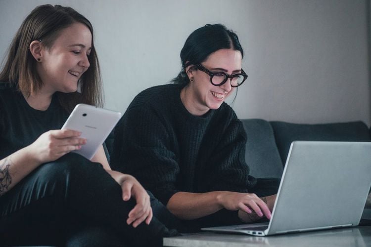 To unge kvinner sitter i en sofa og ser på en pc. Hun ene har et lesebrett i hånden.