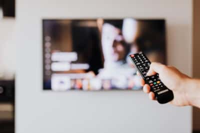 Hånd holder fjernkontroll som peker mot tv på veggen. Tv-bildet er uskarpt i bakgrunnen.