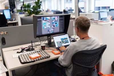 En person sitter ved et arbeidsbord i et åpent kontorlandskap og jobber med bilderedigering på en stor skjerm, mens vedkommende parallelt bruker en bærbar datamaskin. Skrivebordet har flere tekniske hjelpemidler som harddisker, tastatur og kaffekopp.