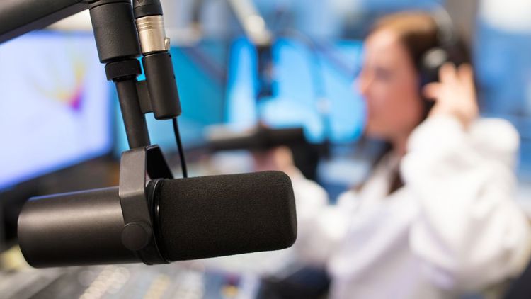 Nærbilde av en radiomikrofon i et studio, med en person med hodetelefoner uskarpt i bakgrunnen. Foto.