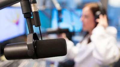 Nærbilde av en radiomikrofon i et studio, med en person med hodetelefoner uskarpt i bakgrunnen. Foto.