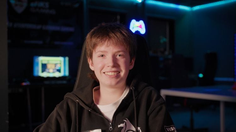 Ung gutt sitter i en gamingstol i et rom med blå belysning og datamaskiner i bakgrunnen. Han smiler og har på seg en hettegenser med logoer.
