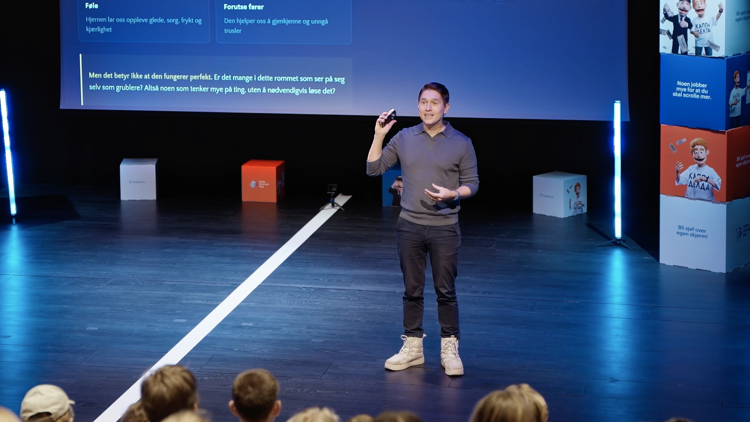 En foredragsholder står på en scene og holder en presentasjon for et publikum. Han holder en liten fjernkontroll i hånden og gestikulerer mens han snakker. Bak ham vises en presentasjon på en stor skjerm med norsk tekst om hjernen og følelser. Scenen er opplyst med blått lys, og publikum sitter foran scenen.