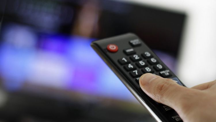 Nærbilde av fjernkontroll. Uskarp tv i bakgrunn.