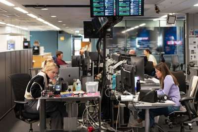 Et åpent kontorlandskap i en redaksjon, der flere personer sitter foran dataskjermer og jobber konsentrert. Skjermer henger fra taket med oversikter og statistikk. Bordene er fulle av teknisk utstyr, kabler og personlige eiendeler. Stemningen virker fokusert og travel.