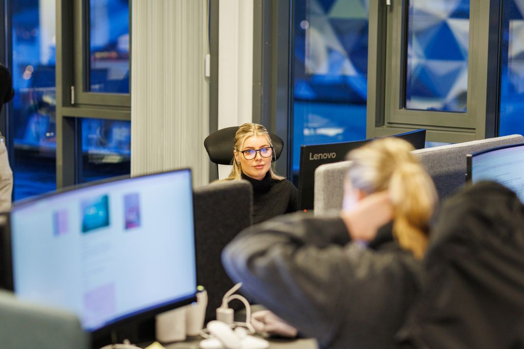 To kvinner i en avisredaksjon sitter ved hver sin arbeidsplass skrått overfor hverandre. Hun lengst fra kamera ser på kollegaen, som bildet viser bakfra. Begge har lyst hår og mørk genser.