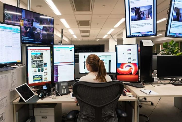 Kvinne sitter ved en pult med ryggen mot kamera. Foran seg og på sidene har hun mange skjermer som viser forskjellige nettaviser og en som viser en tv-sending.