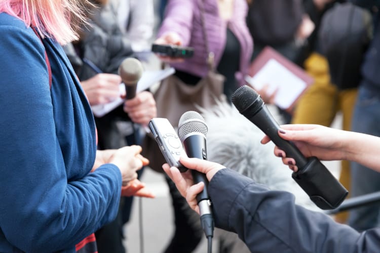 En person blir intervjuet av mange journalister som holder flere mikrofoner foran personen.