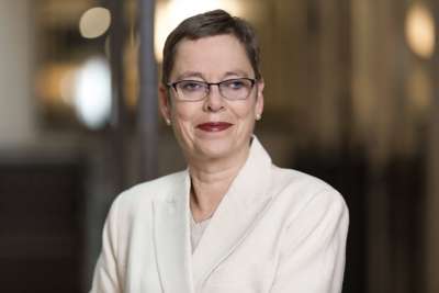 Portrett av Mari Velsand, direktør i Medietilsynet. Hun har kort hår, briller og røde lepper, og er kledd i en lys jakke. Bakgrunnen er uskarp og nøytral.