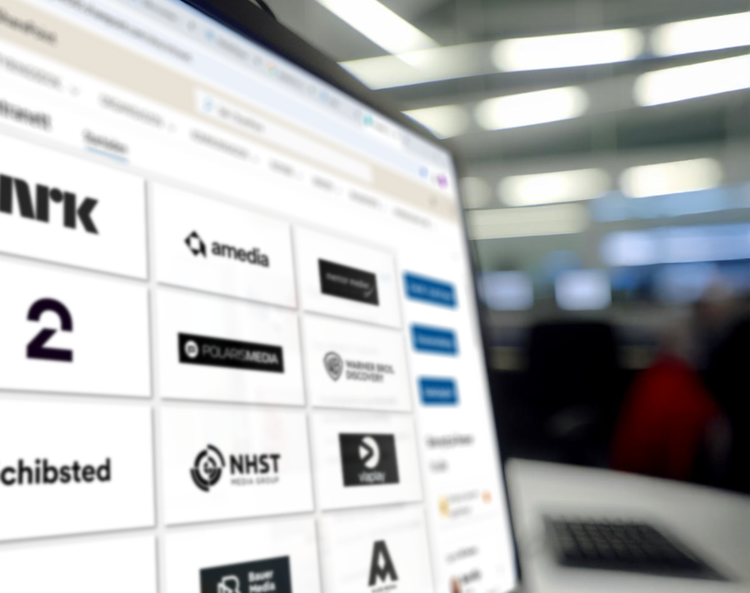 Dataskjerm med oversikt over norske mediehus, inkludert logoene til NRK, TV 2, Schibsted, Amedia, Polaris Media, NHST Media Group, Warner Bros. Discovery og flere. Bildet er tatt i et kontormiljø med uskarp bakgrunn.