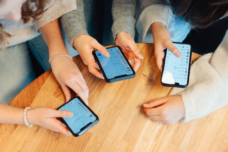 Nærbilde av hender og mobiltelefoner. Tre jenter sitter ved et bord. Vi ser at alle har Snapchat oppe på telefonskjermen.