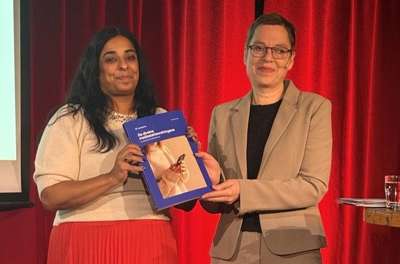 Kultur- og likestillingsminister Lubna Jaffery, og direktør i Medietilsynet, Mari Velsand, står foran et rødt sceneteppe og holder sammen rapporten ‘De direkte mediestøtteordningene’
