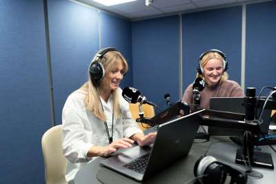 To kvinner sitter i et radiostudio og smiler. Begge har hodetelefoner på og pc-skjerm foran seg. Den ene snakker i en mikrofon.