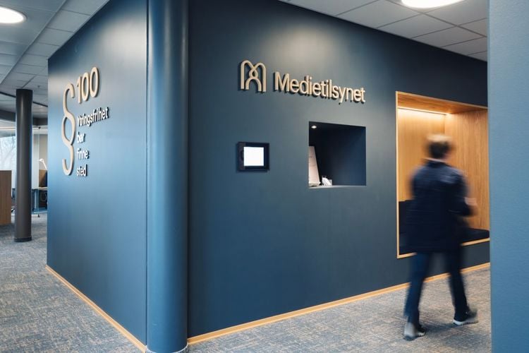 Bilde fra Medietilsynets kontor. En person beveger seg bort fra kamera med ryggen til. På en vegg står det Medietilsynet, rundt hjørnet står det paragraf hundre, ytringsfrihet bør finne sted.