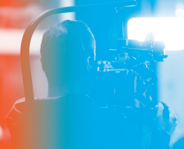 En videojournalist sett bakfra står med et stort videokamera på skuldra.