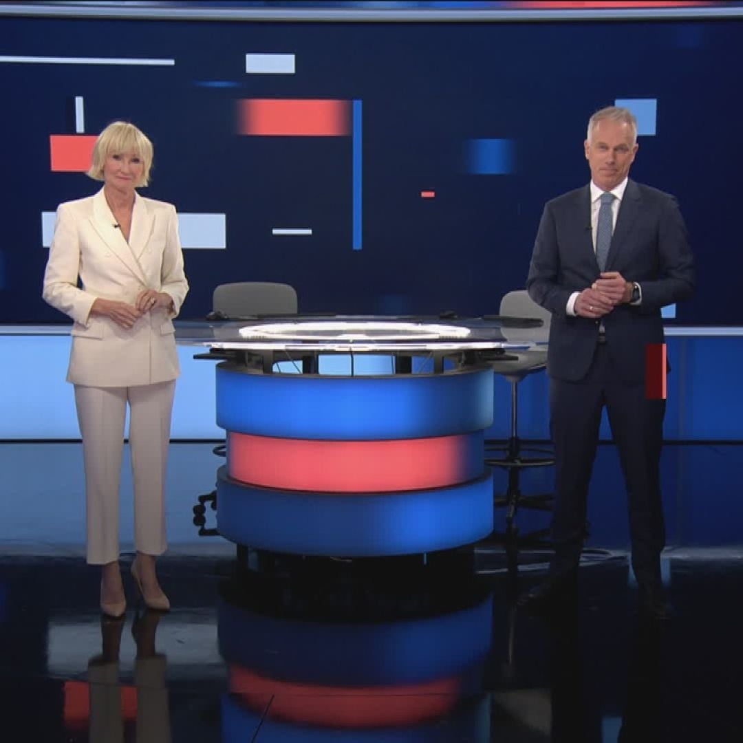 To nyhetsankere st&aring;r p&aring; hver sin side av et rundt nyhetsbord med bl&aring;tt og r&oslash;dt lys, foran en stor skjerm med abstrakte bl&aring;, r&oslash;de og hvite grafiske elementer. Foto.