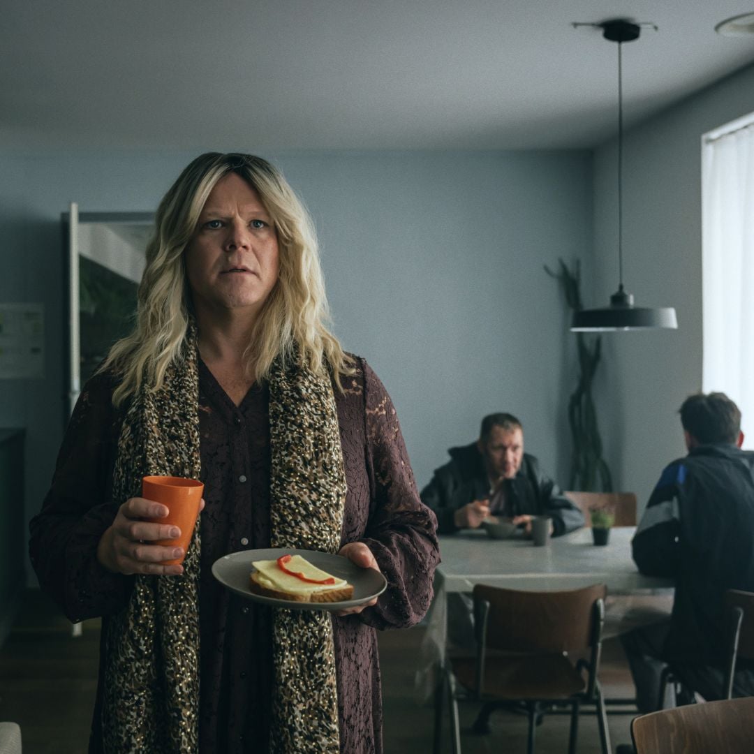 En kvinne med langt blondt h&aring;r holder en tallerken med en br&oslash;dskive og et oransje krus, st&aring;ende i et rom med to personer som sitter ved et bord i bakgrunnen. Rommet har n&oslash;ytrale farger, en hengelampe over bordet og et vindu som slipper inn naturlig lys. Foto.