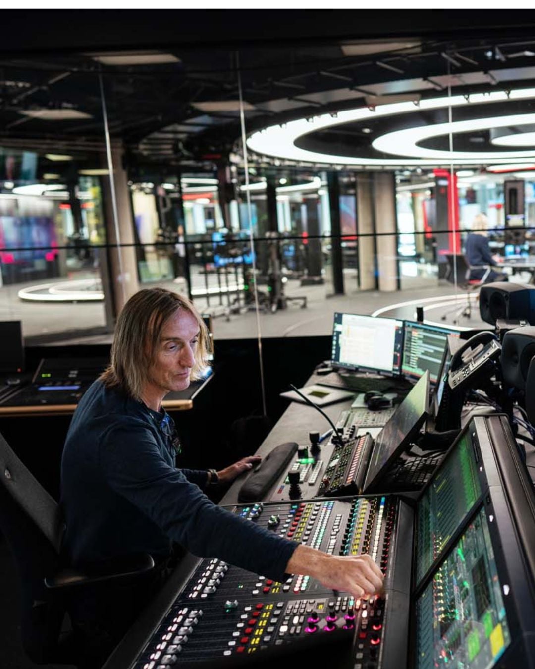 Ein person sit ved ein stor miksepult med fleire skjermar i eit kontrollrom for radio- eller TV-produksjon.