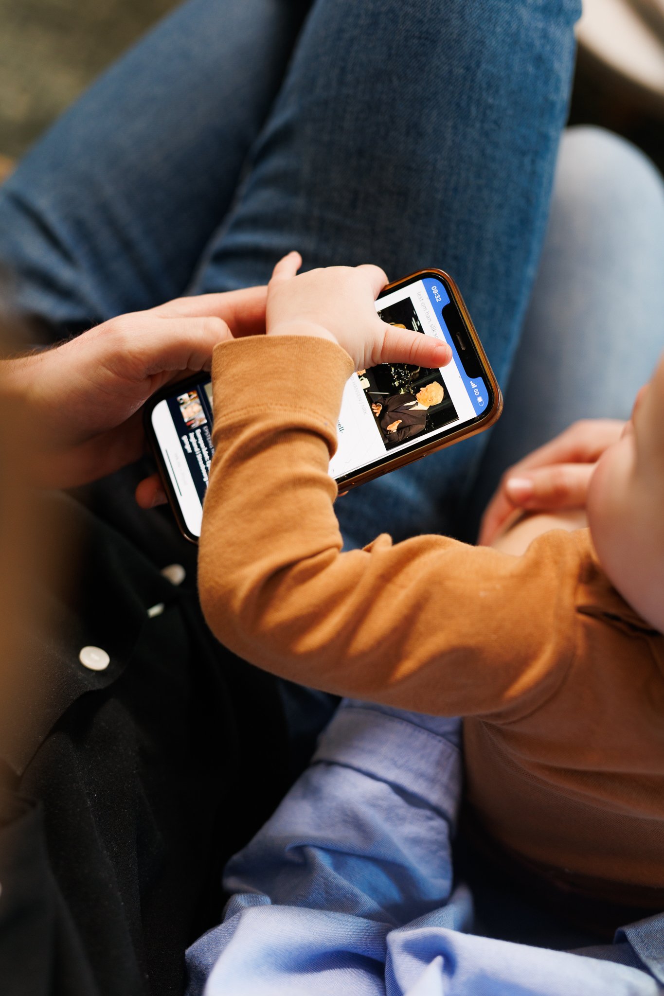 Barn sitter p&aring; fanget til en voksen og bruker en mobiltelefon til &aring; se p&aring; innhold p&aring; skjermen.