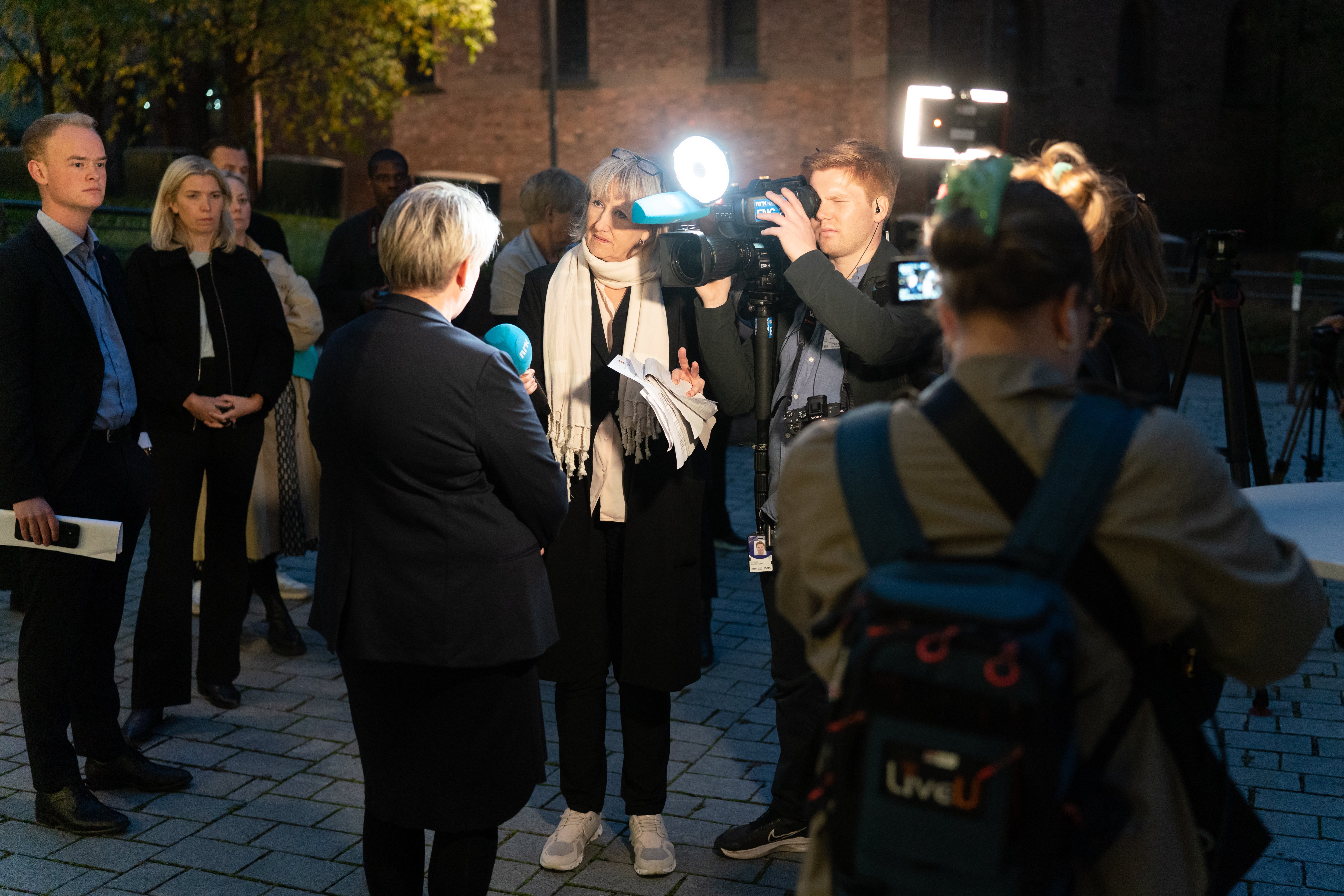 Journalist intervjuer person foran kamera og lysrigg under pressekonferanse utend&oslash;rs.