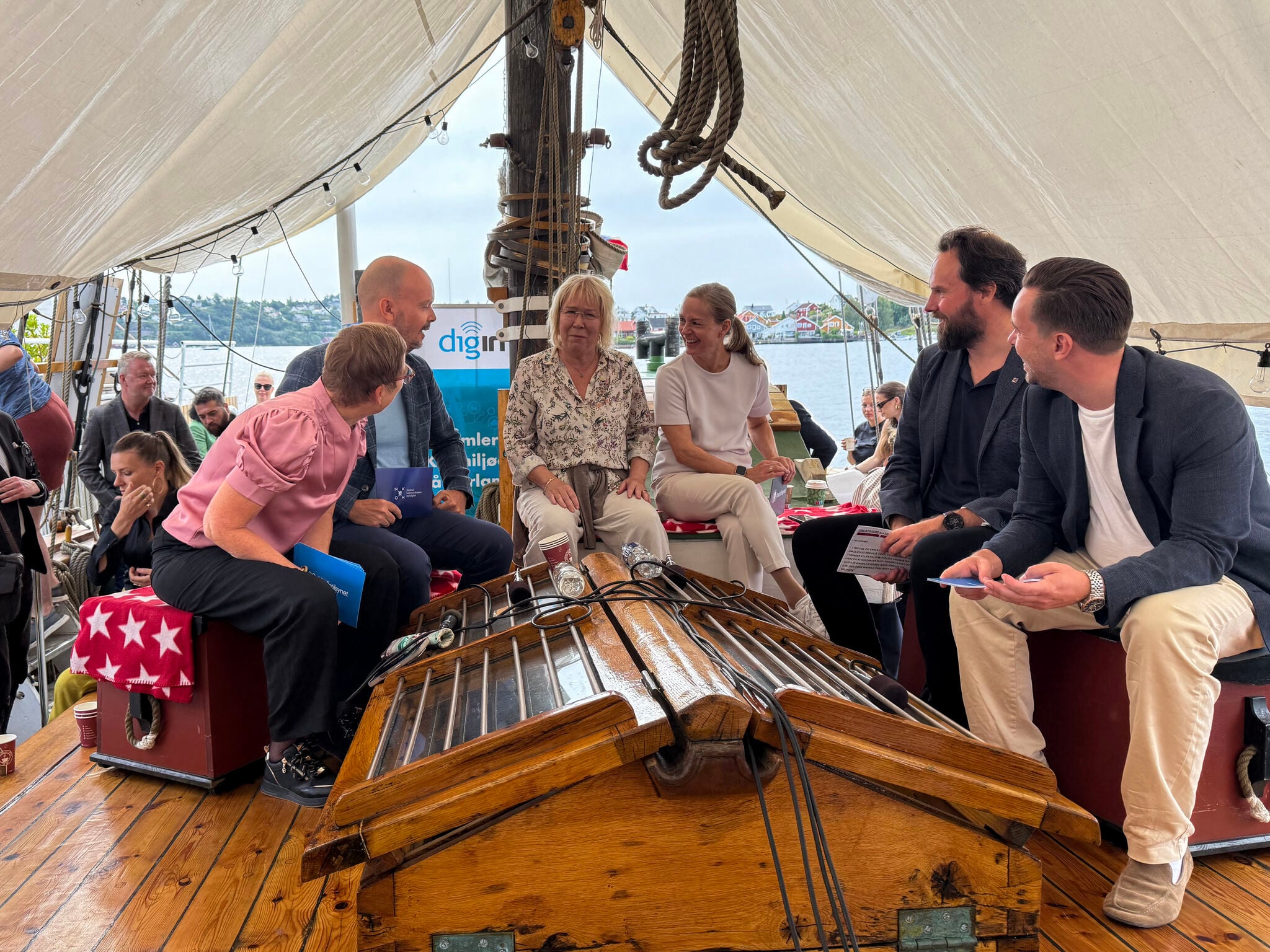 Panelsamtale om bord i en seilb&aring;t under Arendalsuka. Seks personer sitter i en sirkel og diskuterer, med mikrofoner p&aring; bordet. Havn og bebyggelse er synlig i bakgrunnen.