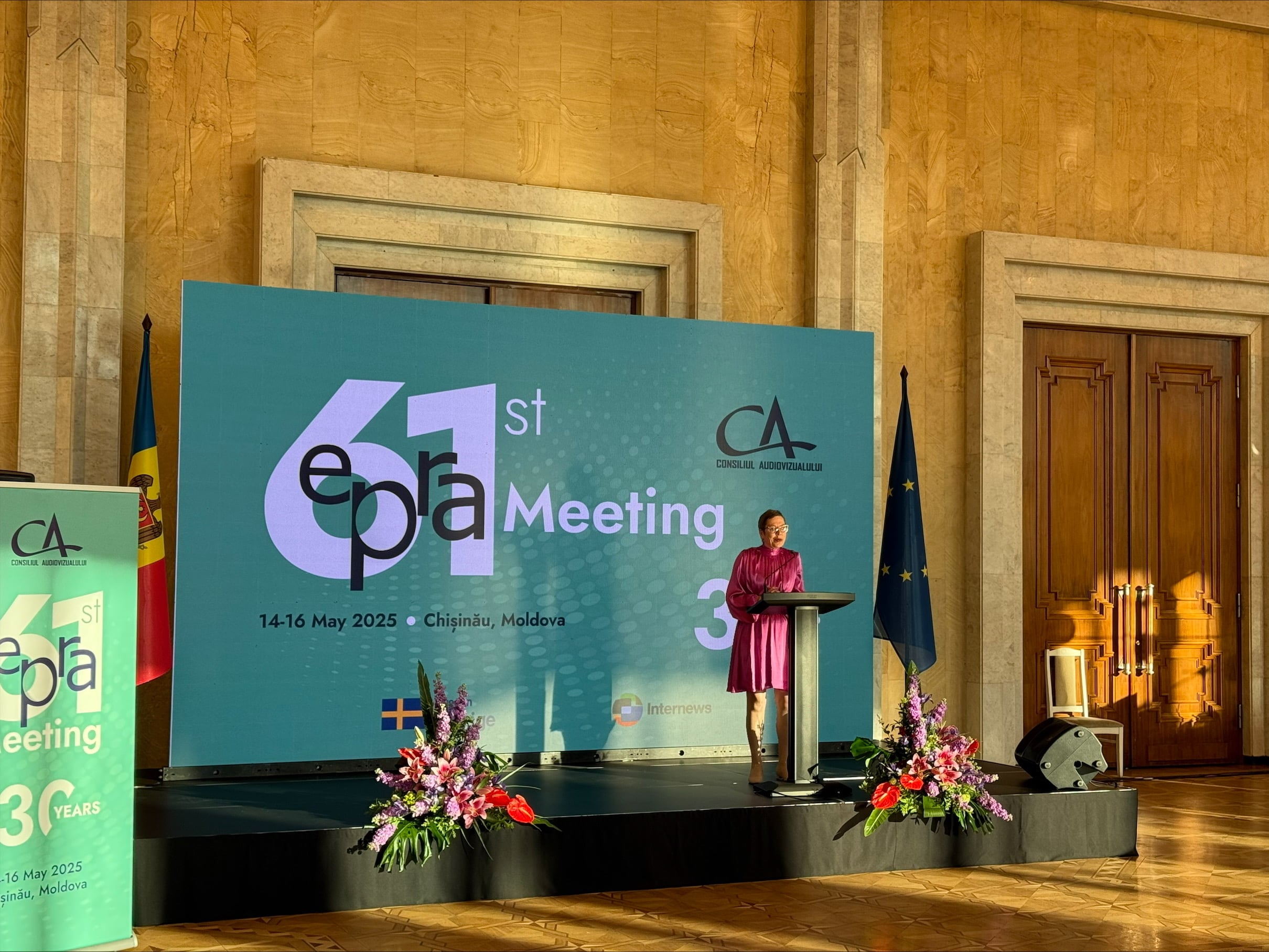 En kvinne holder innlegg fra en talerstol under det 61. EPRA-m&oslash;tet i Chișinău i Moldova. P&aring; scenen st&aring;r blomsterdekorasjoner og flagg fra Moldova og EU.