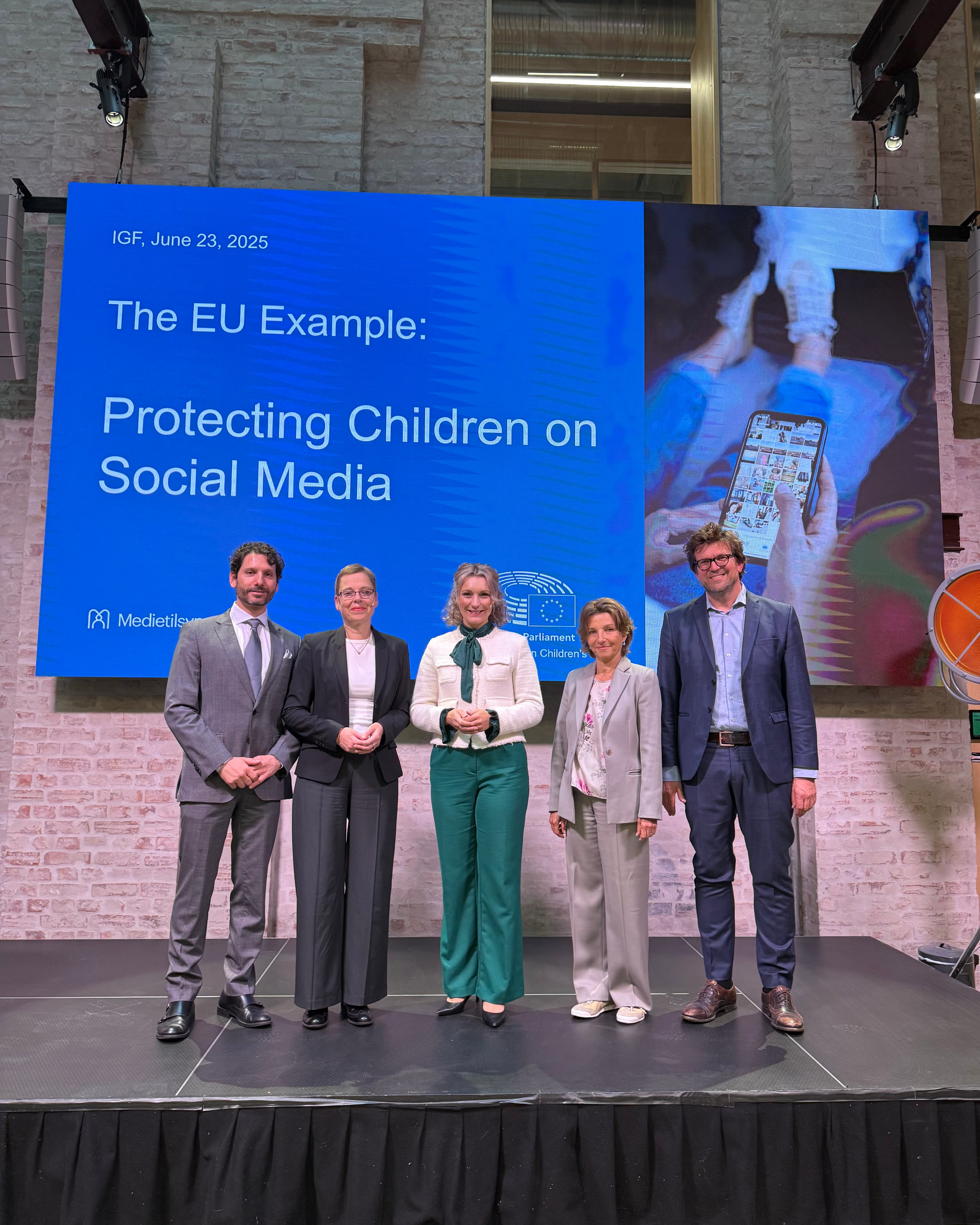 Fem personer st&aring;r p&aring; en scene foran en stor skjerm med teksten &laquo;The EU Example: Protecting Children on Social Media&raquo; under Internet Governance Forum 2025.