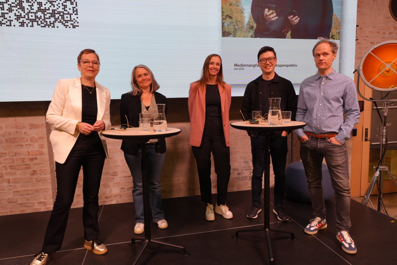 Fem personer st&aring;r p&aring; en scene foran en presentasjon med tittelen &laquo;Mediemangfold i et bruksperspektiv&raquo;. Foran dem st&aring;r to h&oslash;ye bord med vannkarafler og glass.
