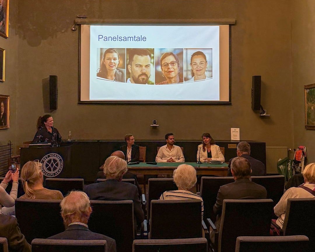 Panelsamtale i en sal med publikum. Fire personer sitter p&aring; scenen, og en moderator st&aring;r ved en talerstol. P&aring; lerretet bak vises tittelen &laquo;Panelsamtale&raquo; og portretter av deltakerne.
