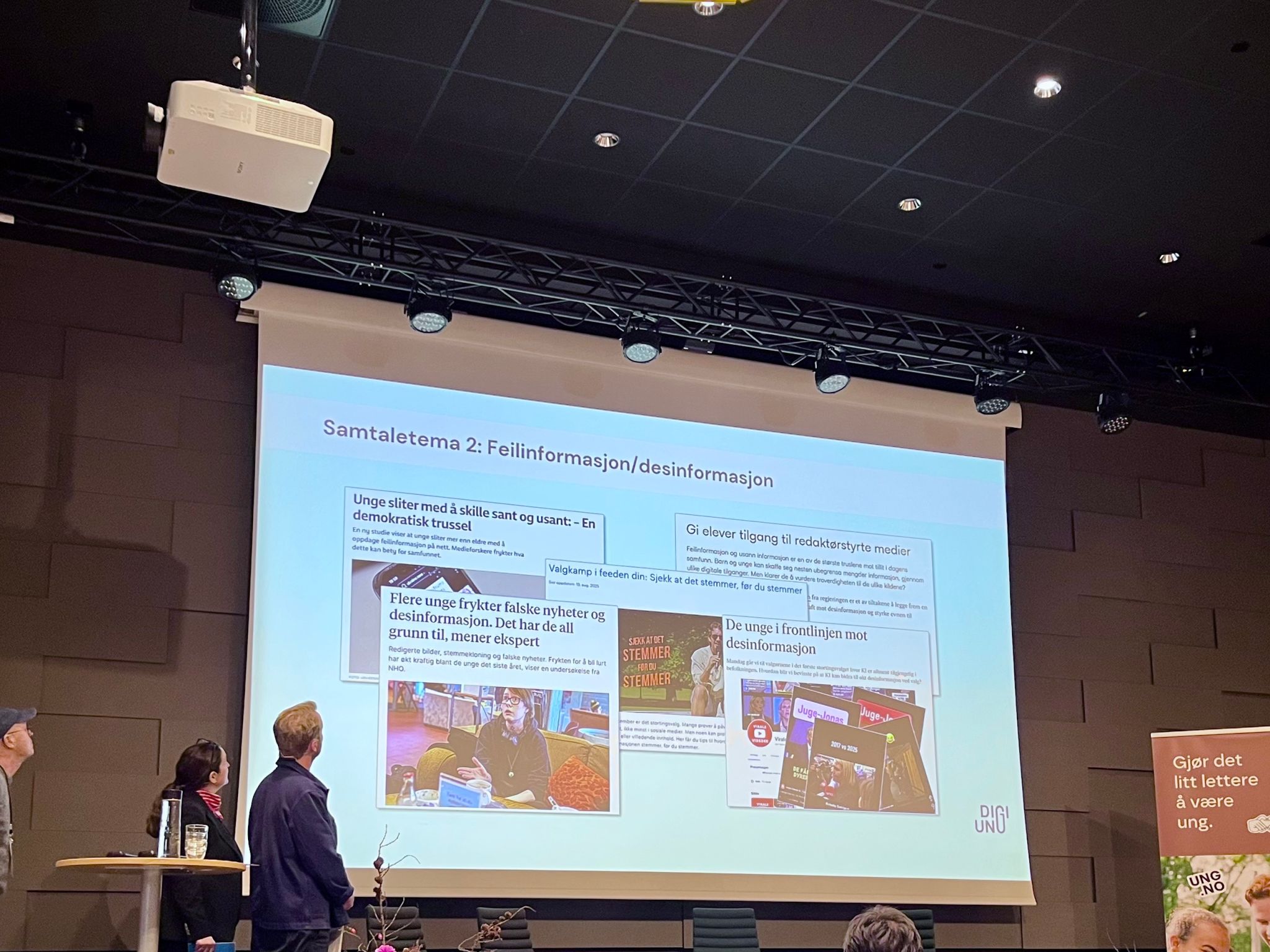 To personer st&aring;r p&aring; en scene og ser p&aring; en stor skjerm med en presentasjon om feilinformasjon og desinformasjon. P&aring; lysbildet vises overskrifter og eksempler fra mediesaker, mens publikum sitter foran scenen.
