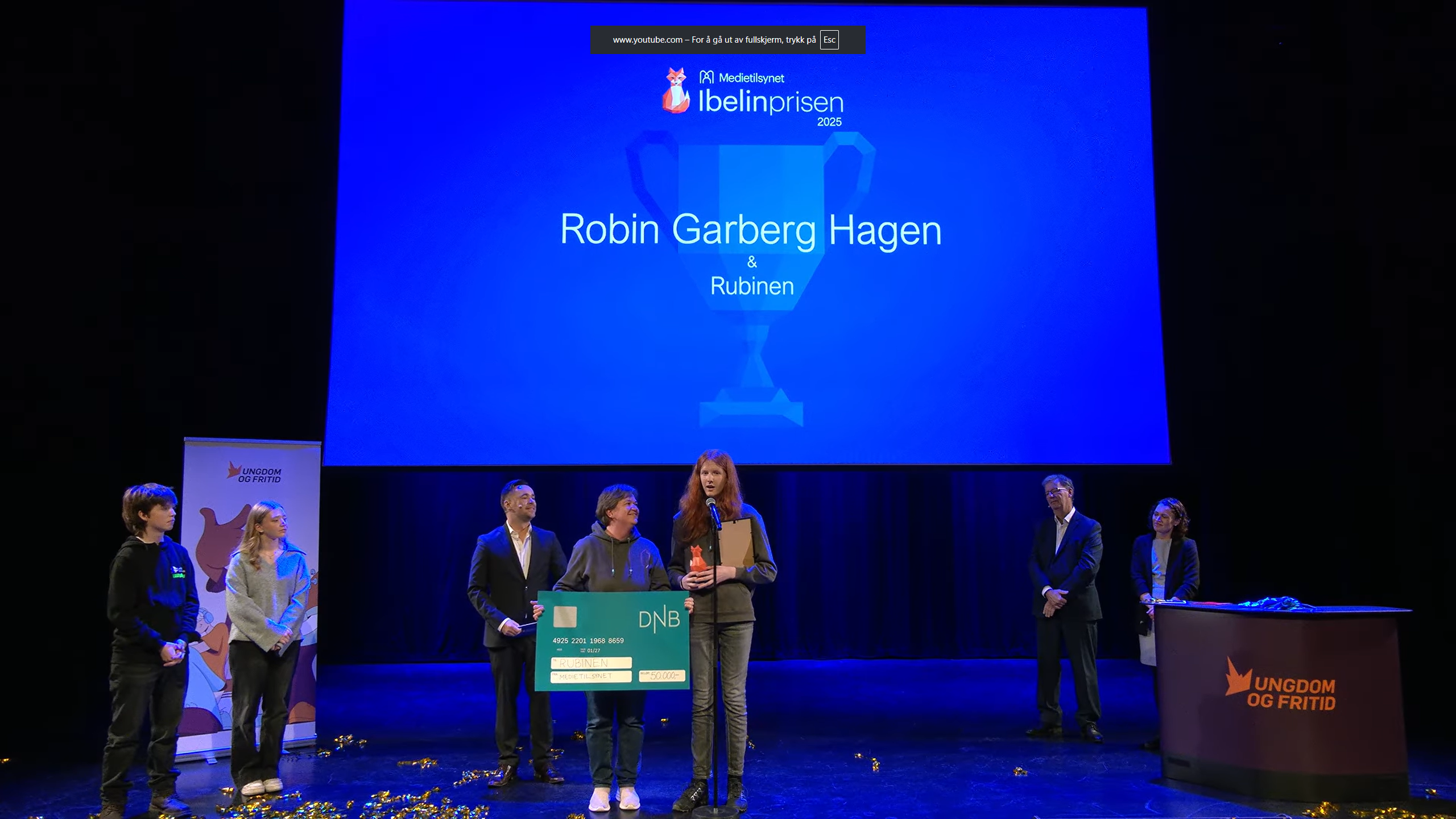 Flere personer st&aring;r p&aring; en scene under utdeling av Ibelinprisen 2025. En person holder tale ved mikrofon, mens andre st&aring;r ved siden av med en stor sjekk. P&aring; skjermen bak vises navnet &laquo;Robin Garberg Hagen & Rubinen&raquo;.